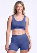 A woman with long blonde hair smiles while wearing the Woolx Aubrey Lounge Bra in Cloudy Bay, paired with matching high-waisted boxer shorts, against a plain white background.