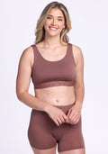 A smiling blonde woman wears the Woolx Aubrey Lounge Bra in Nutmeg with high-waisted shorts, standing against a plain white background.