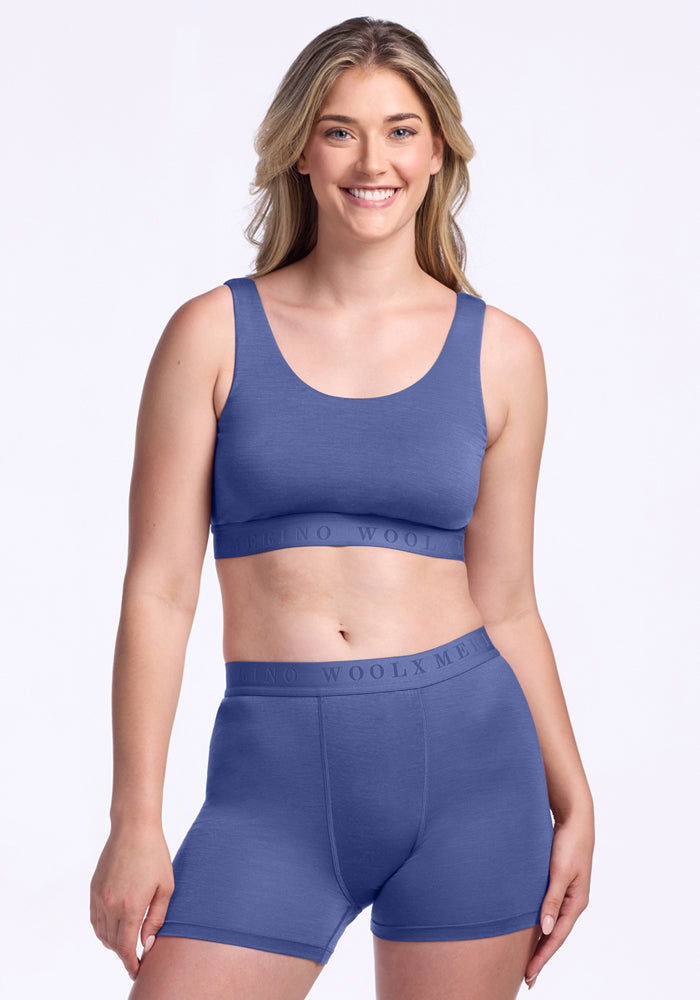 A woman with long blonde hair smiles while wearing the Woolx Aubrey Lounge Bra in Cloudy Bay, paired with matching high-waisted boxer shorts, against a plain white background.