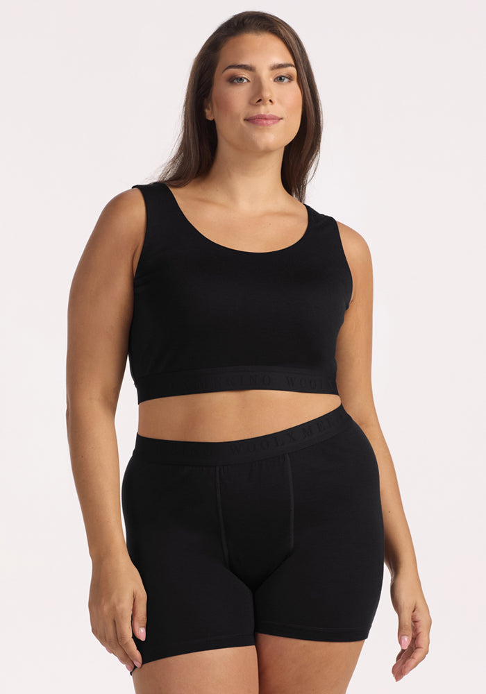 A woman with long brown hair wears the Woolx Aubrey Lounge Bra in black, featuring a logo band, and matching high-waisted shorts. She stands against a plain light background, facing the camera with a neutral expression.