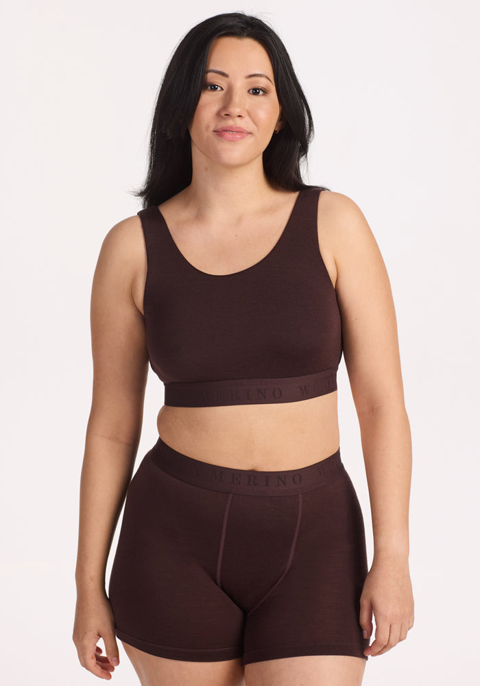 A woman with long dark hair stands against a plain background, wearing the Woolx Aubrey Lounge Bra in Chocolate Plum and matching high-waisted shorts. She faces forward and smiles softly.