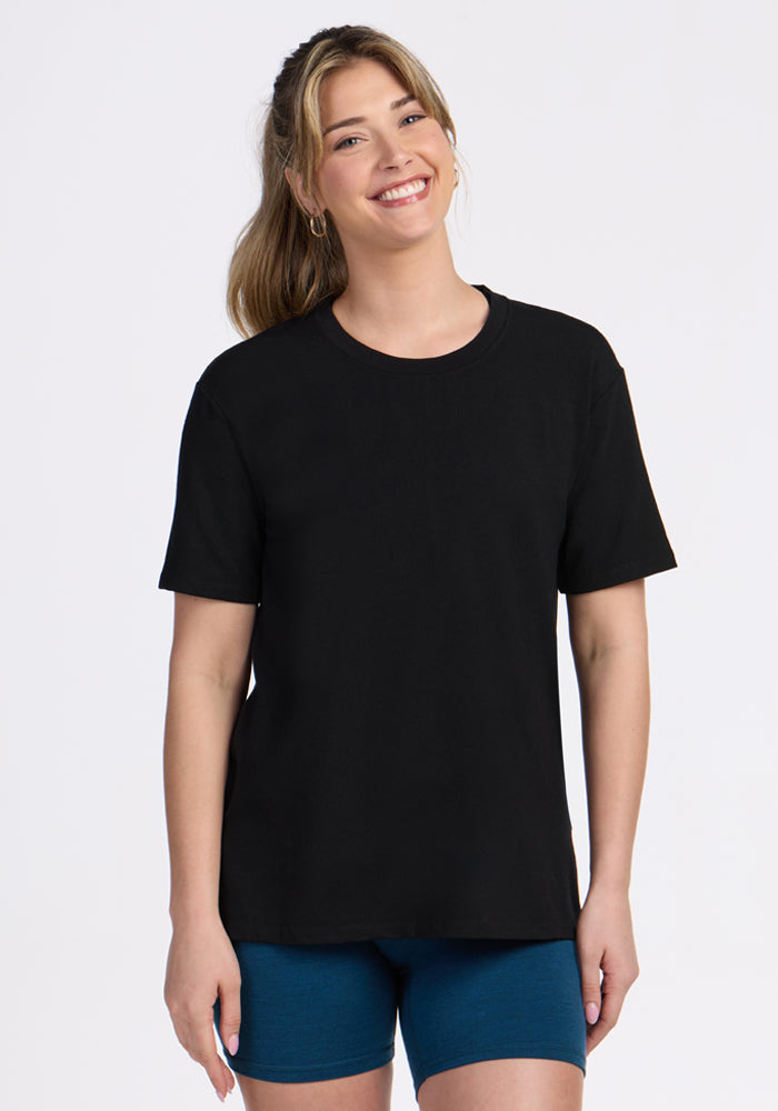 A woman with long, light brown hair smiles in a relaxed fit Woolx Audrey Tee - Black and blue shorts, standing against a simple white background. | Ariana is 5'11", wearing a size S