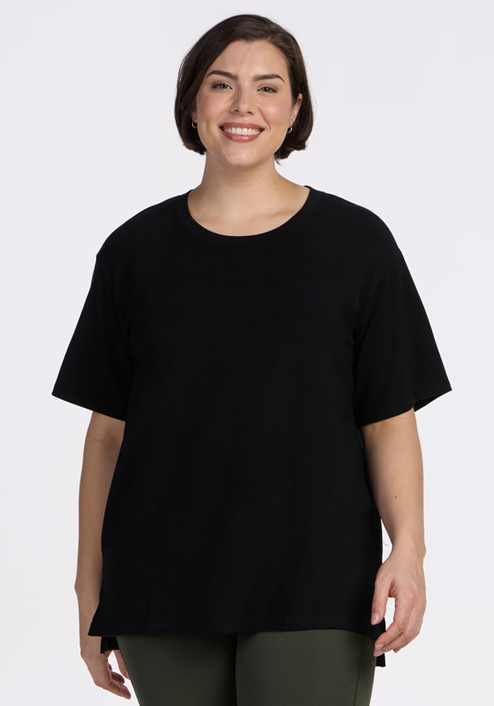 A woman with short brown hair smiles while wearing the Woolx Audrey Tee in black and dark green pants, standing against a plain white background. | Juliana is 5'10", wearing a size XL