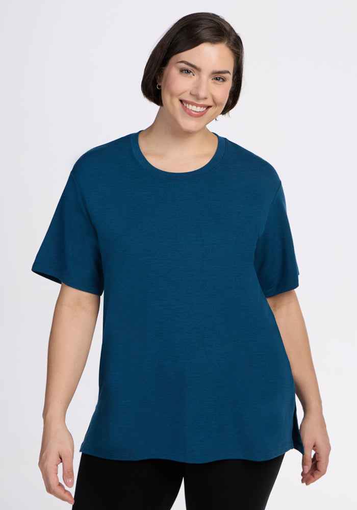 A smiling woman with short dark hair wears the Woolx Audrey Tee in Majolica Blue and black pants, standing against a plain white background. | Juliana is 5'10", wearing a size XL