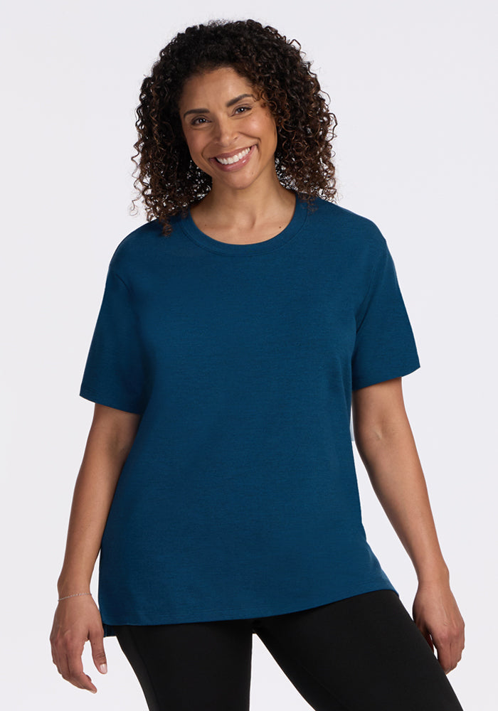 A woman with curly hair smiles while wearing the Woolx Audrey Tee in Majolica Blue and black pants, standing against a plain white background. | Kisha is 5’8”, wearing a size M