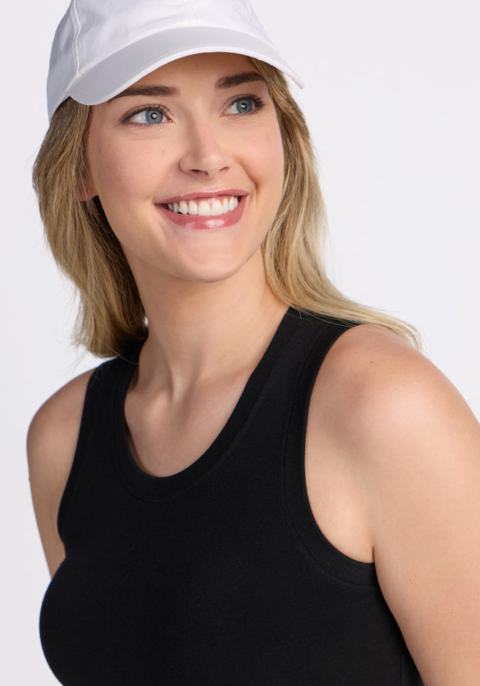 A woman with long blonde hair smiles and looks to the side, wearing a white baseball cap and the Woolx Ava Tank Top in black against a light background.