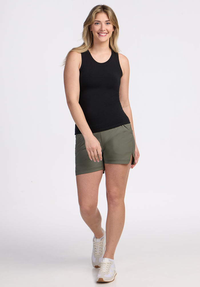 A smiling woman with long blonde hair models the Woolx Ava Tank Top in black, paired with olive green shorts and white sneakers, standing against a plain white background.