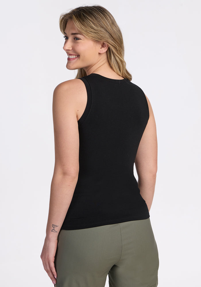 A person with light hair, shown from the back and smiling to the side, wears a Woolx Ava Tank Top - Black and olive green pants, revealing a small tattoo on their left forearm.
