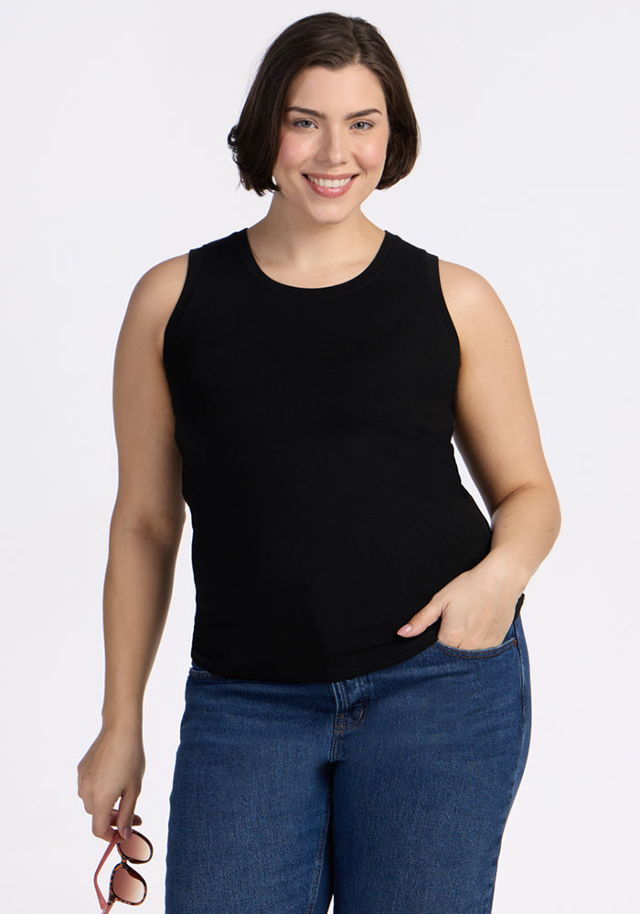 A woman with short brown hair, wearing the Woolx Ava Tank Top in black and blue jeans, smiles at the camera against a plain white background, holding sunglasses in her left hand. | Juliana is 5'10", wearing a size XL