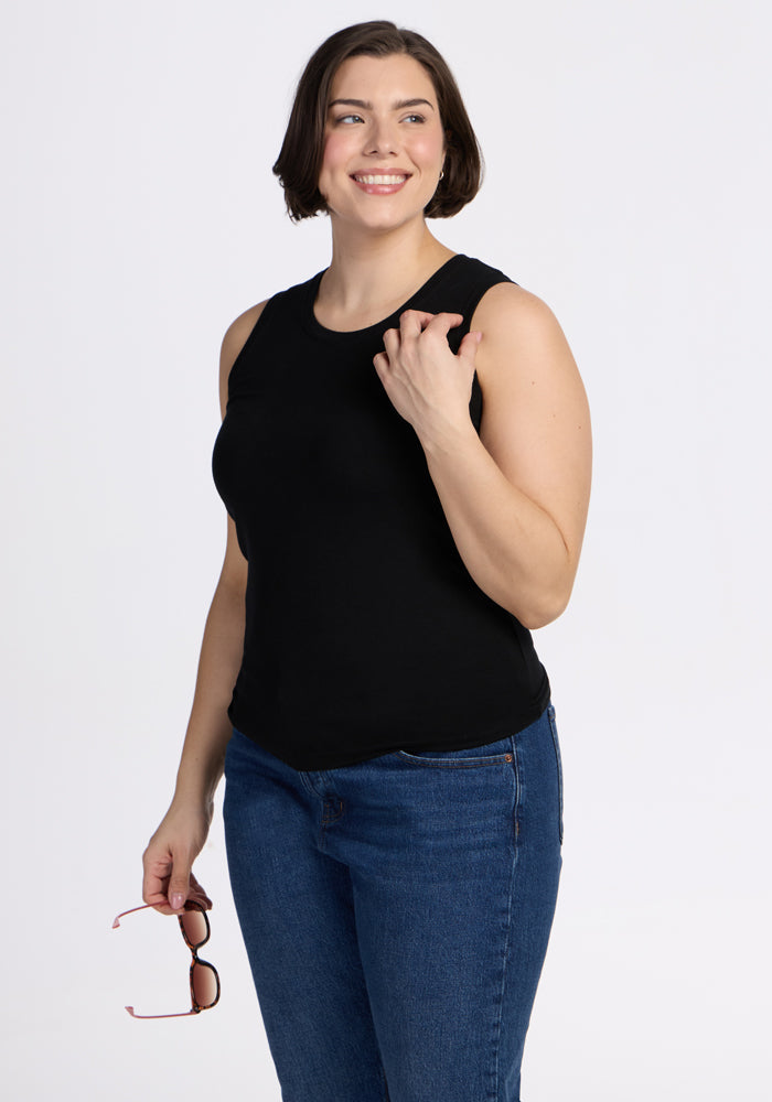 A woman with short brown hair smiles against a white background, wearing the Woolx Ava Tank Top in black and blue jeans, holding sunglasses in her left hand.