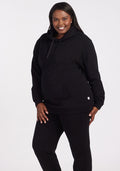 A woman smiles against a white background, wearing the Woolx Avery Hoodie in black with matching black pants, one hand in her pocket and the other by her side. | Le'Quita is 5'11", wearing a size XL