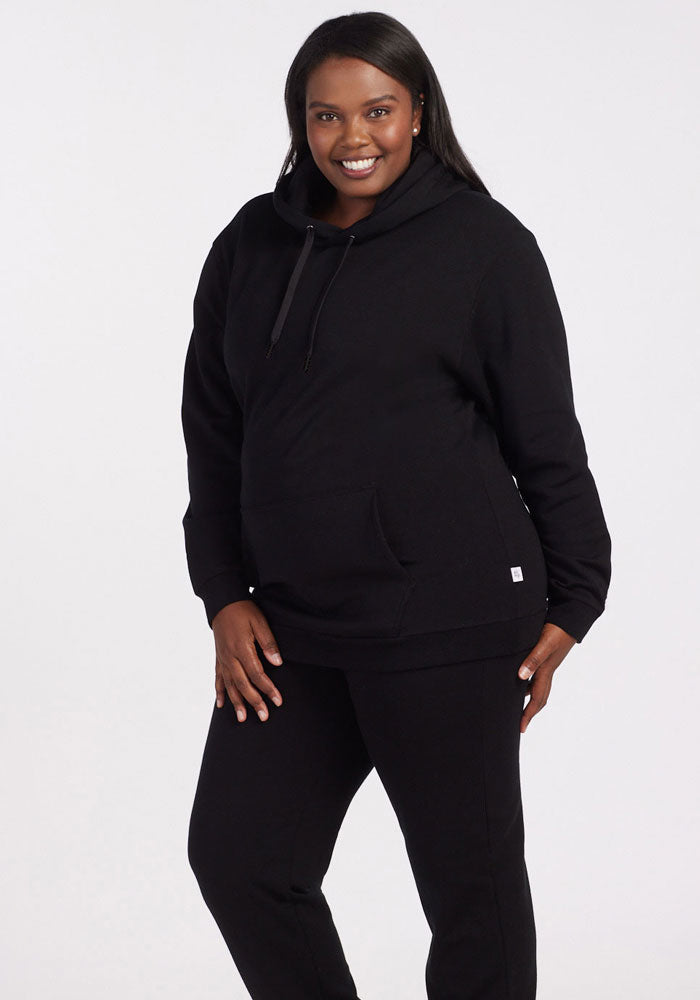 A woman smiles against a white background, wearing the Woolx Avery Hoodie in black with matching black pants, one hand in her pocket and the other by her side. | Le'Quita is 5'11", wearing a size XL