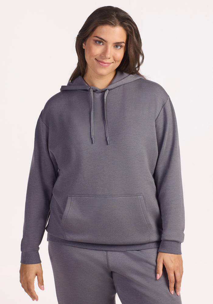A woman smiles softly against a light background, wearing the Woolx Avery Hoodie in Arctic Dusk and matching sweatpants, with her right hand relaxed by her side.