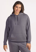 A woman smiles softly against a light background, wearing the Woolx Avery Hoodie in Arctic Dusk and matching sweatpants, with her right hand relaxed by her side.