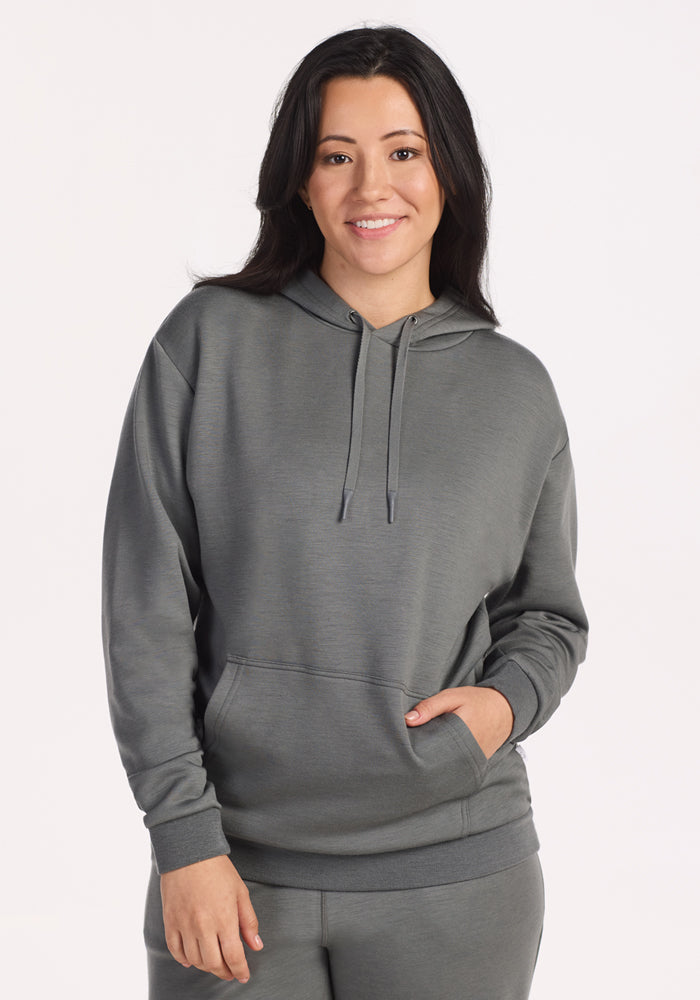 A woman with long dark hair smiles in the Woolx Avery Hoodie • Final Sale - Sedona Sage, featuring a front pocket and matching gray pants. This cozy set is made from soft, odor-resistant fabric for lasting comfort against a plain, light background.