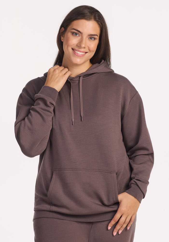 A woman with long brown hair smiles, wearing the Woolx Avery Hoodie in Plum Truffle (final sale), featuring a cozy front pocket and drawstrings, standing against a plain light background.