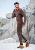 A man stands on snow before large rocks, smiling and raising a hand near his face. He wears Woolx Backcountry Leggings in Bourbon Oak—a fitted merino wool base layer—paired with tan hiking boots, ideal for staying warm outdoors.