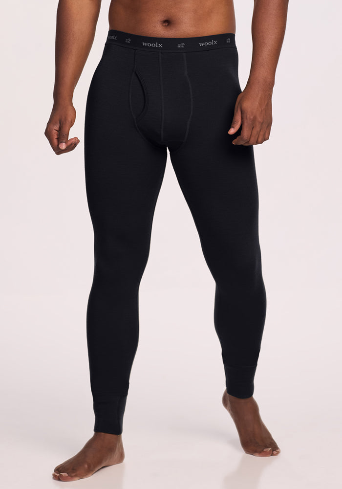A person stands barefoot against a plain light background, dressed in black Woolx Backcountry Leggings with a visible "woolx" waistband, showcasing fitted Merino wool gear perfect for cold weather adventures.