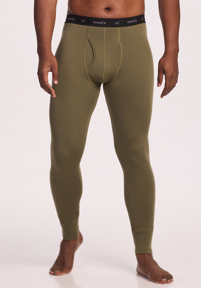 A man stands barefoot on a plain, light background wearing Woolx Backcountry Leggings in Winter Moss, featuring a black "woolx" waistband.