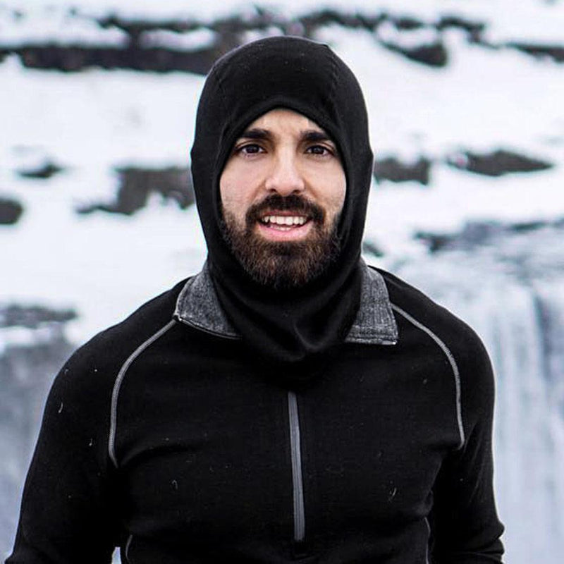 An outdoor enthusiast with a beard is smiling at the camera, wearing a black Woolx Balaclava Head Warmer and a black jacket made of merino wool. He is standing outdoors in a snowy landscape with snow-covered rocks in the background.