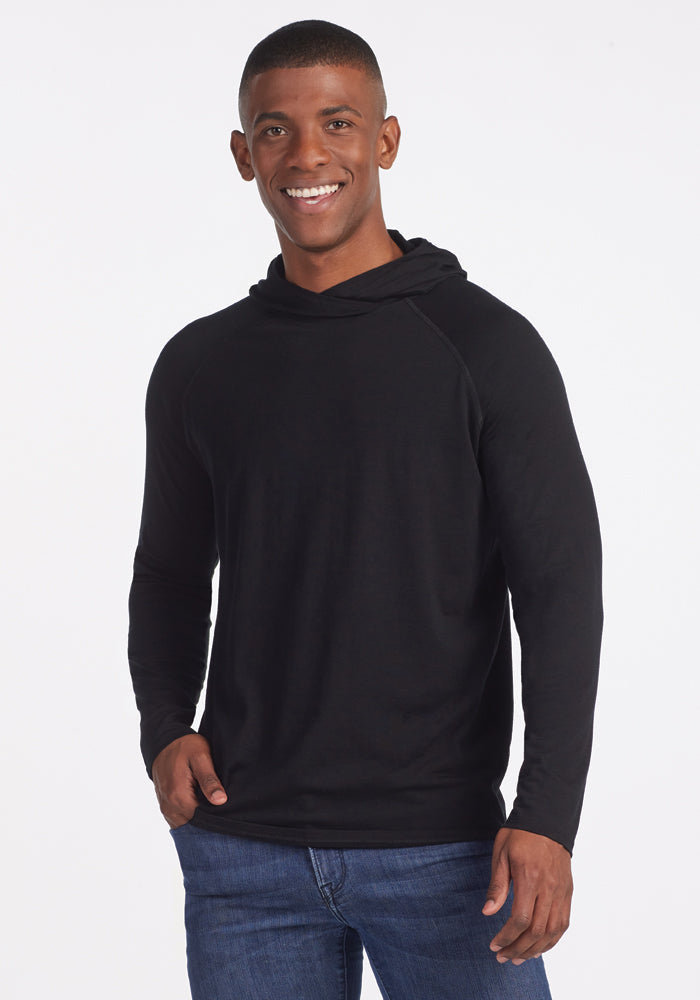 A person wearing a Base Camp Hoodie in black by Woolx and blue jeans smiles at the camera against a white background. | Trell is 6’2”, wearing a size M