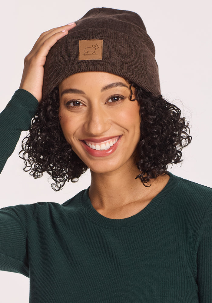 A smiling woman wears the Woolx Baylor Beanie - Bracken, featuring a small logo patch and crafted from Australian Merino Wool, paired with a green ribbed long-sleeve shirt as she touches the beanie against a light background.