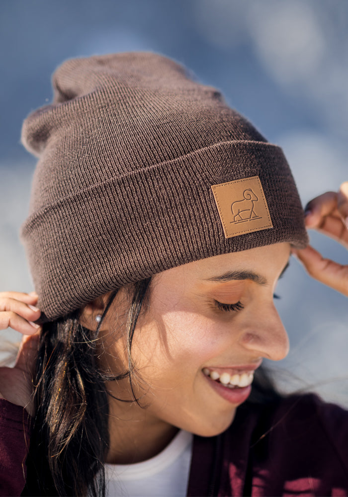 A smiling person adjusts the Woolx Baylor Beanie - Bracken, a brown merino wool hat with an animal logo leather patch, while standing outdoors in soft natural light.