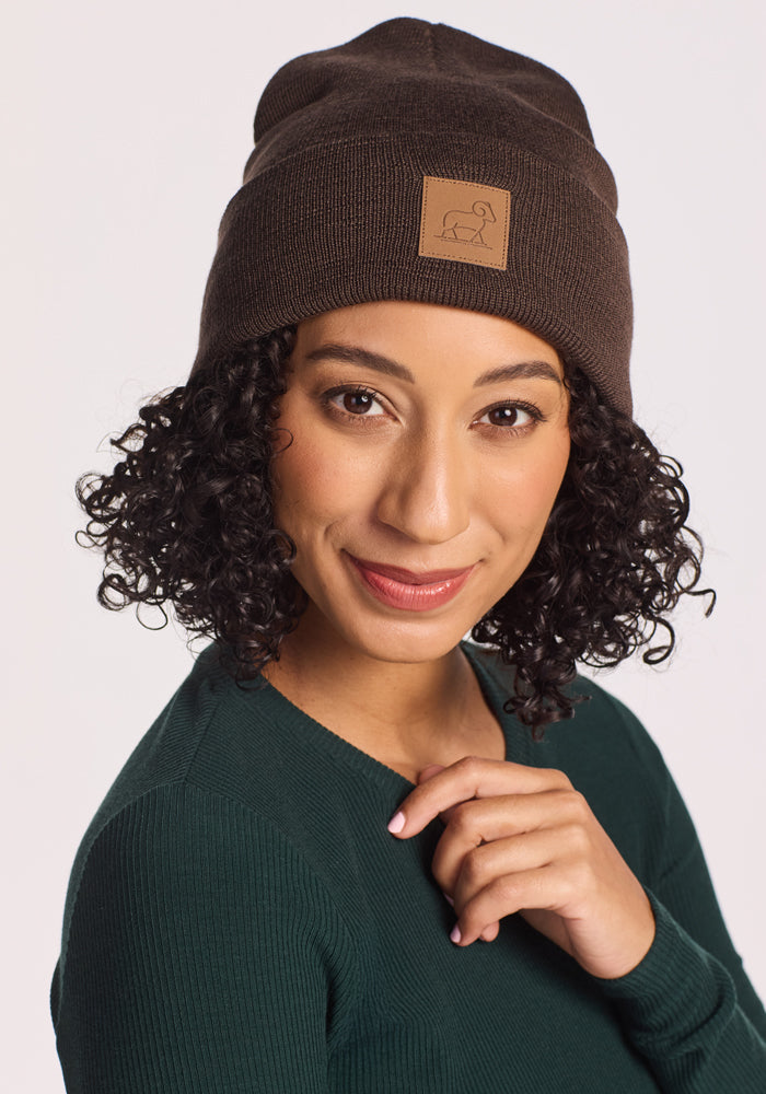 A woman with curly dark hair wears the Woolx Baylor Beanie • Final Sale - Bracken, featuring a square patch, and a green sweater. She smiles gently, touching the edge of her beanie against a plain white background.
