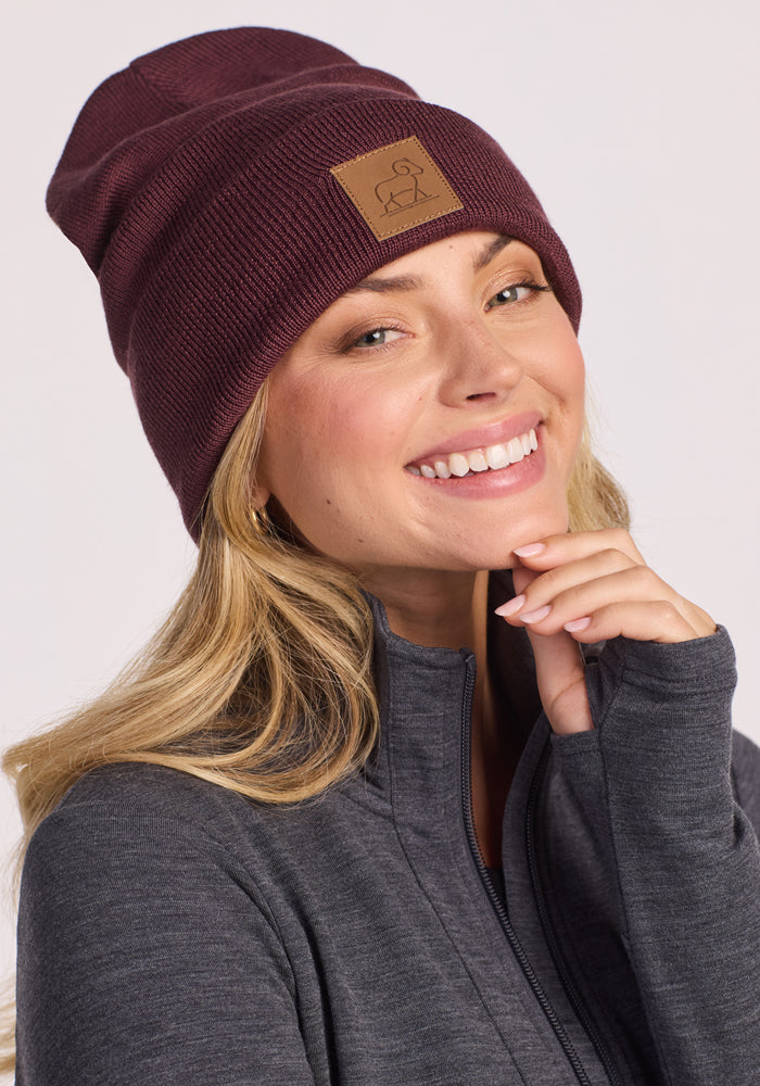 A smiling woman with long blonde hair wears the Woolx Baylor Beanie in Grape Harvest and a dark gray zip-up jacket. She touches her chin with one hand and looks directly at the camera.
