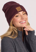 A smiling woman with long blonde hair wears the Woolx Baylor Beanie in Grape Harvest and a dark gray zip-up jacket. She touches her chin with one hand and looks directly at the camera.