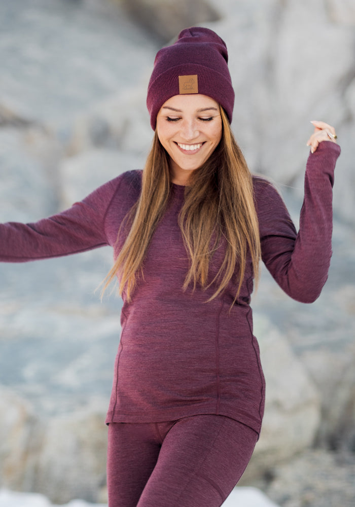A woman with long brown hair smiles outdoors on a snowy day, wearing the Woolx Baylor Beanie in Grape Harvest. She appears joyful, dressed in a matching maroon outfit, with a blurred rocky background behind her.