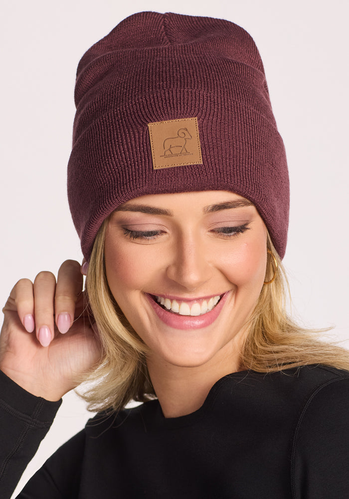 A smiling person in a black top wears the Woolx Baylor Beanie - Grape Harvest, a maroon merino wool hat with a tan patch featuring a simple animal logo, holding its edge with one hand.