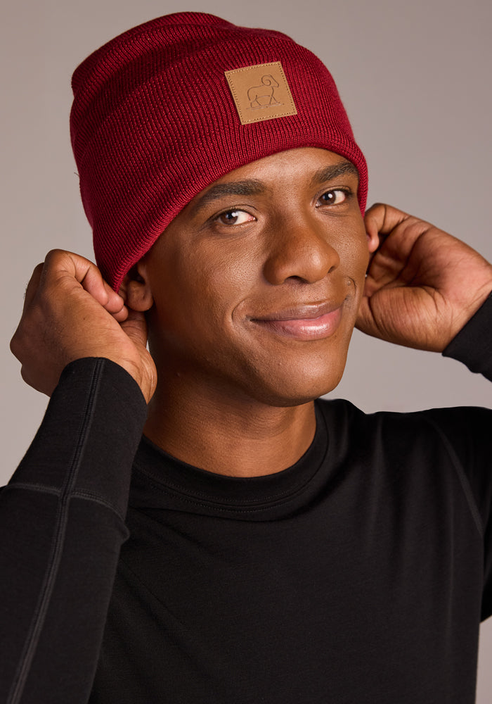 A person wearing the Woolx Baylor Beanie - Rhubarb, featuring a brown patch, and a black long-sleeve shirt smiles softly while adjusting the cozy hat with both hands against a neutral background.