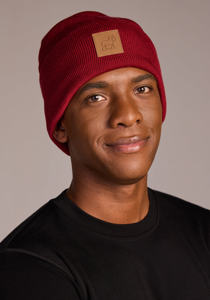 A person wearing the Woolx Baylor Beanie in Rhubarb, featuring a brown patch, pairs it with a black shirt and smiles softly against a neutral background.