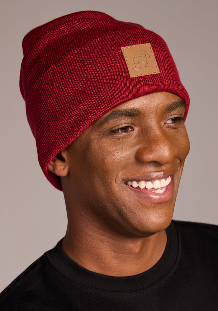 A smiling person in a Woolx Baylor Beanie - Rhubarb, featuring a tan front patch and black shirt, poses against a neutral background.