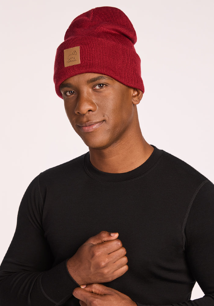 A man models the Woolx Baylor Beanie - Rhubarb, featuring a brown patch, paired with a black long-sleeve shirt against a light backdrop. His subtle smile and hand on chest highlight this cozy winter hat.