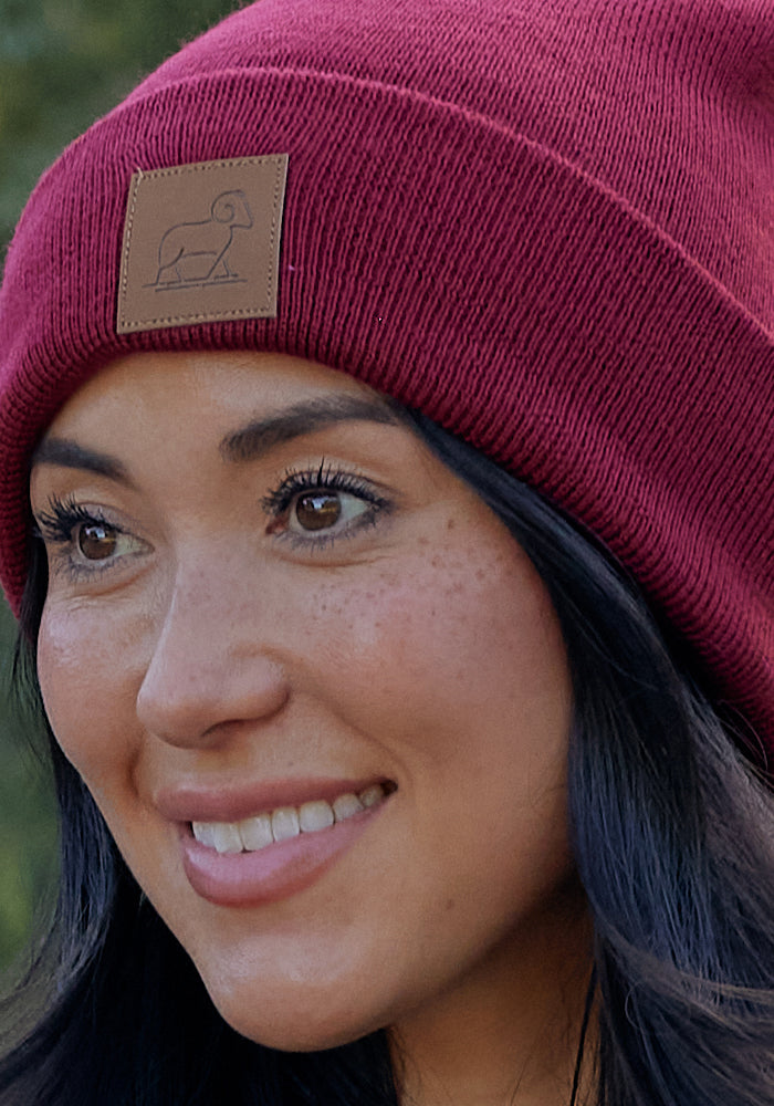 A woman with long dark hair smiles while wearing the Woolx Baylor Beanie in Sweet Cream, accented by a brown patch featuring a simple ram logo—a cozy winter hat perfect for chilly days.