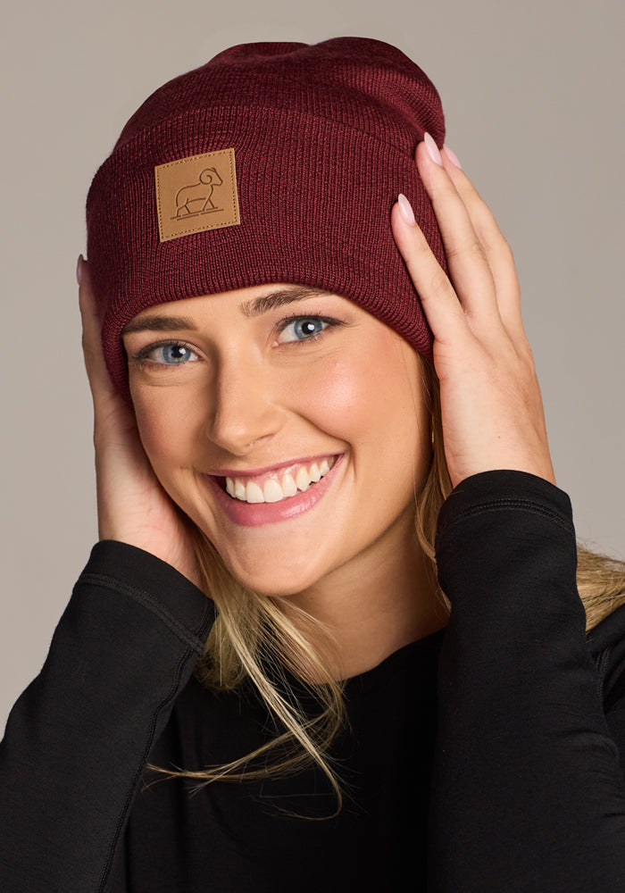 A smiling woman in a black top wears the Woolx Baylor Beanie in Toasted Garnet, a warm winter hat made from Australian merino wool. She holds the beanie’s sides with both hands, showing its front rectangular patch.