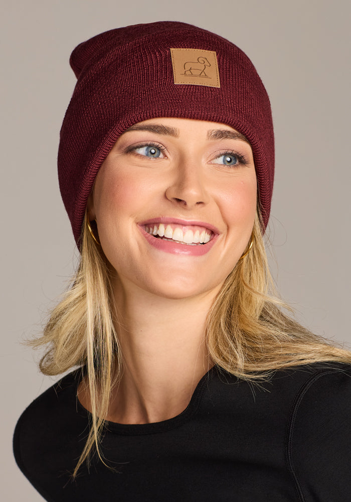 A smiling blonde woman wears the Woolx Baylor Beanie in Toasted Garnet, featuring a brown patch, paired with a black top against a plain light gray background.