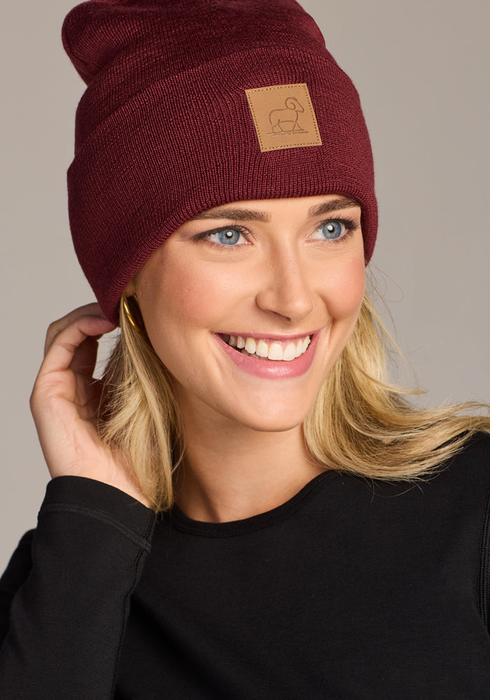 A smiling blonde woman wears the Woolx Baylor Beanie in Toasted Garnet, paired with a black top. She looks to the side, touching the beanie featuring a brown patch—a perfect warm winter hat.