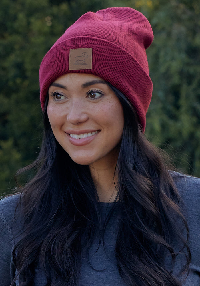A woman with long dark hair is smiling outdoors amid green foliage, wearing a Woolx Baylor Beanie in Toasted Garnet and a gray top.
