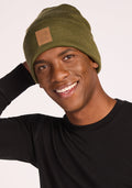A smiling person wears the Woolx Baylor Beanie in Winter Moss and a black long-sleeve shirt, posing with one hand behind their head against a plain light background.