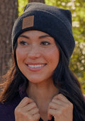 A woman wearing the Woolx Baylor Beanie in Carbon Black and a purple sweater smiles outdoors in a wooded area, sunlight filtering through the trees.