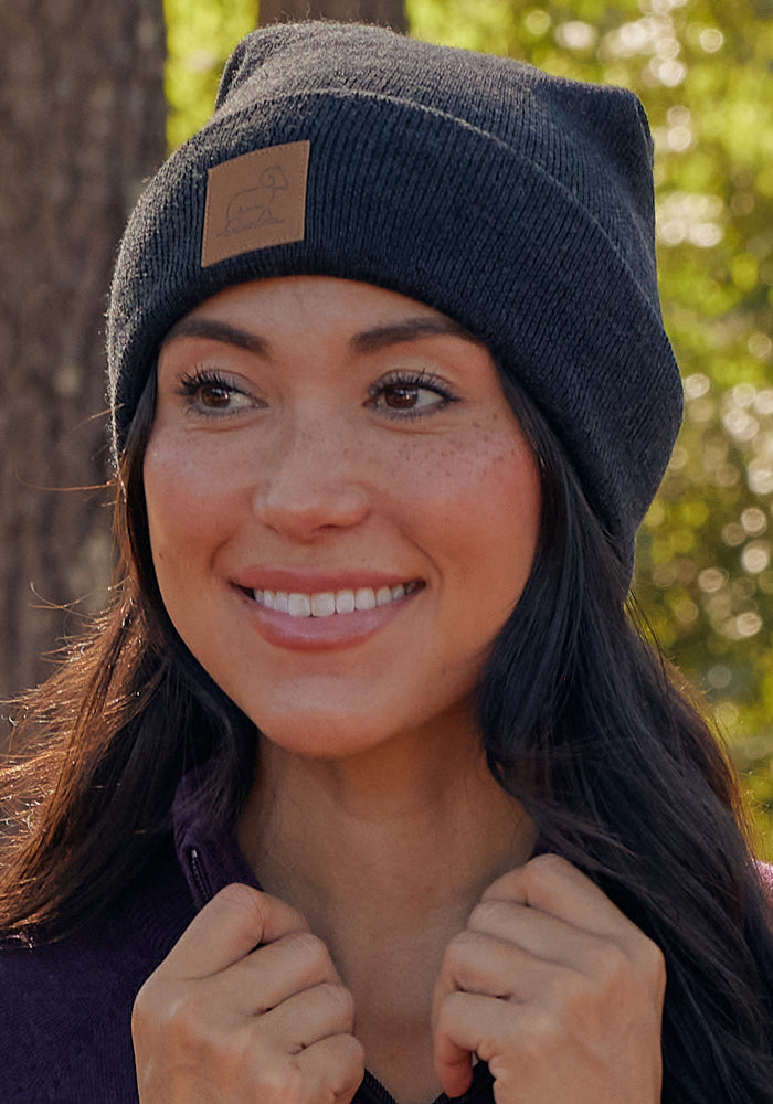 A woman wearing the Woolx Baylor Beanie in Carbon Black and a purple sweater smiles outdoors in a wooded area, sunlight filtering through the trees.