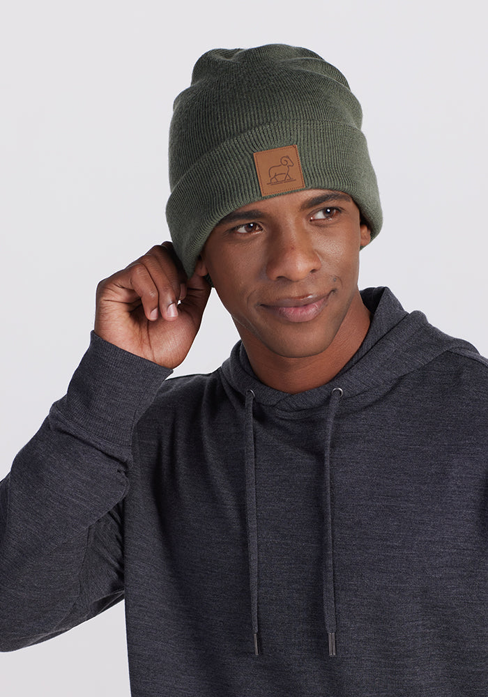 A person wearing a dark gray hoodie adjusts their Woolx Baylor Beanie in Spruce Heather. This beanie, crafted from soft Australian Merino wool, features a brown patch with an animal silhouette. The individual smiles slightly against a neutral background.