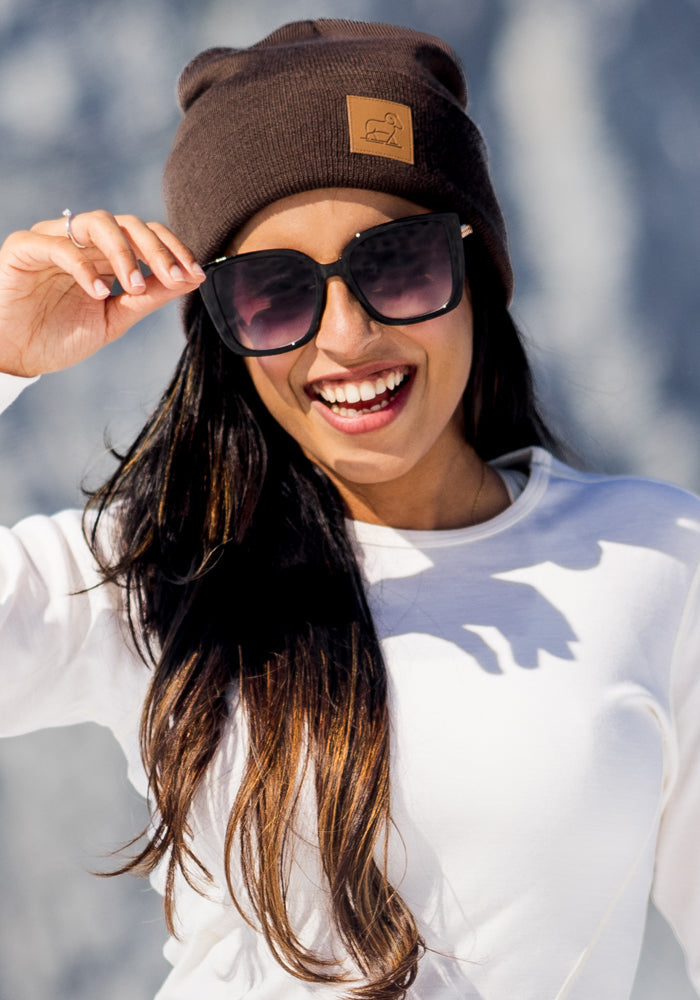 A woman wearing the Woolx Baylor Beanie in Bracken, black sunglasses, and a white long-sleeve shirt stands outdoors in bright sunlight, holding her sunglasses with one hand and smiling.
