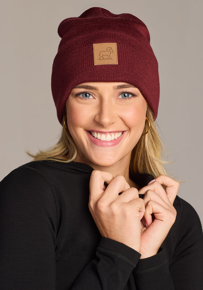 A smiling blonde woman wears the Woolx Baylor Beanie in Toasted Garnet, featuring a stitched patch, along with a black long-sleeve top. She holds her collar with both hands and poses against a neutral background.