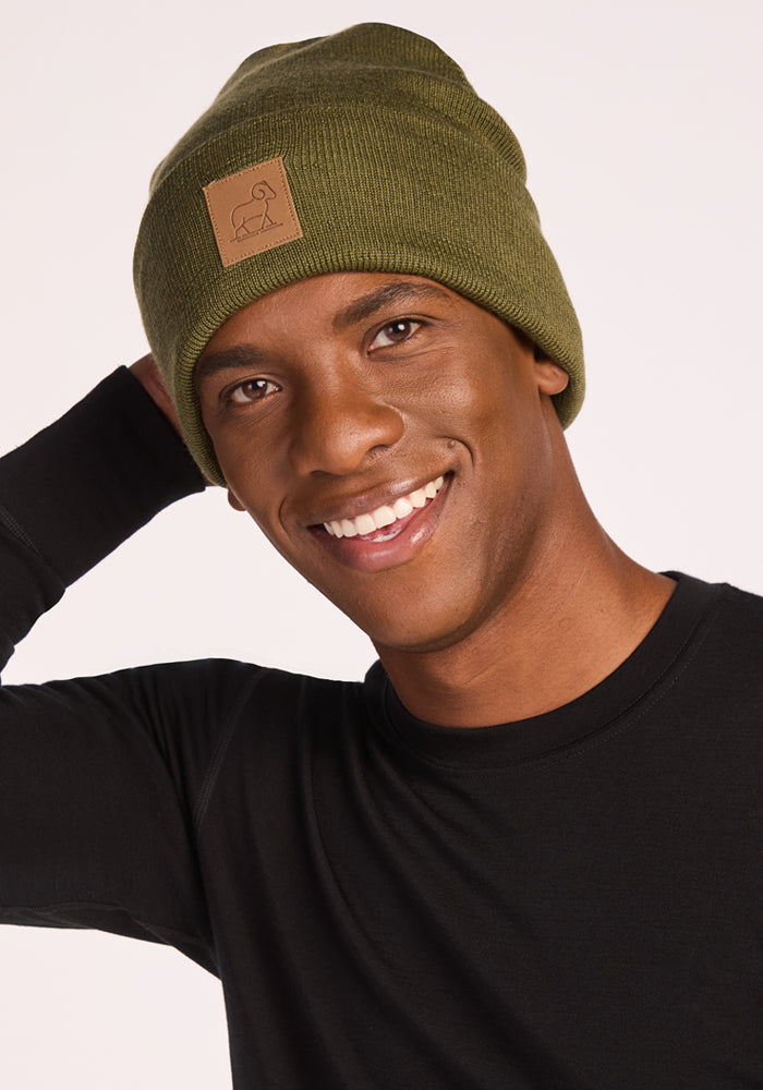 A smiling person wears the Woolx Baylor Beanie in Winter Moss and a black long-sleeve shirt, posing with one hand behind their head against a plain light background.