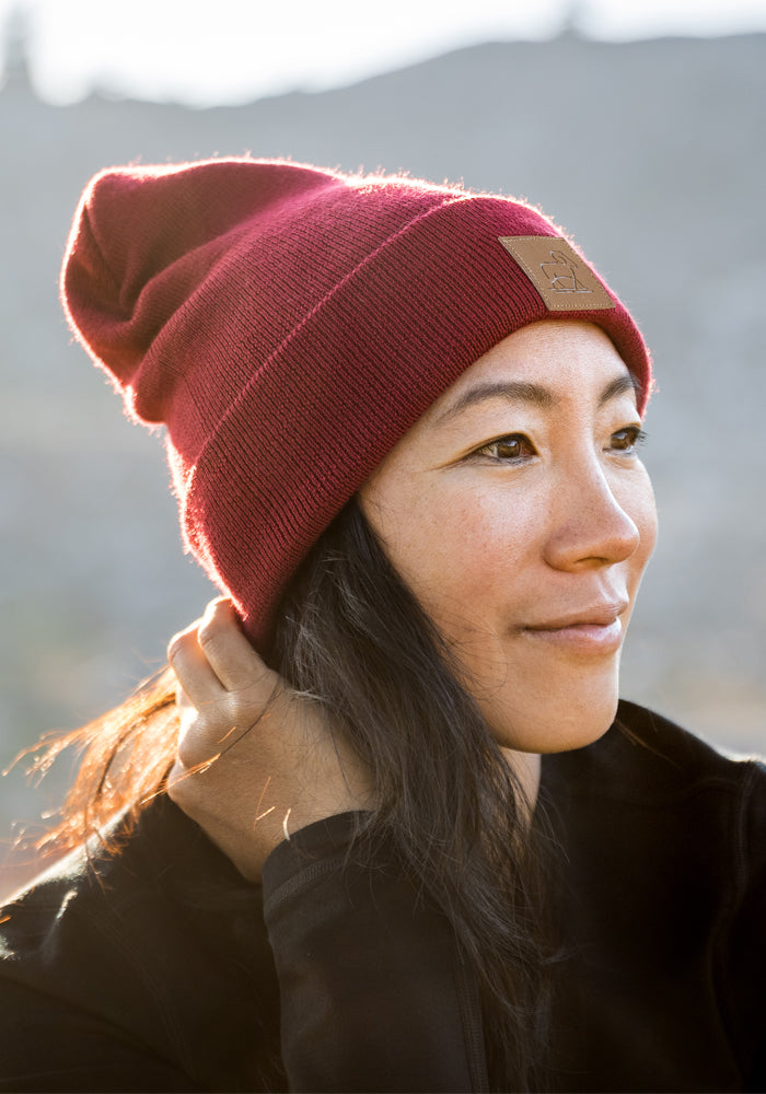 A woman wearing the Woolx Baylor Beanie in Rhubarb and a black jacket smiles softly while gazing into the distance, set against a blurred outdoor background.