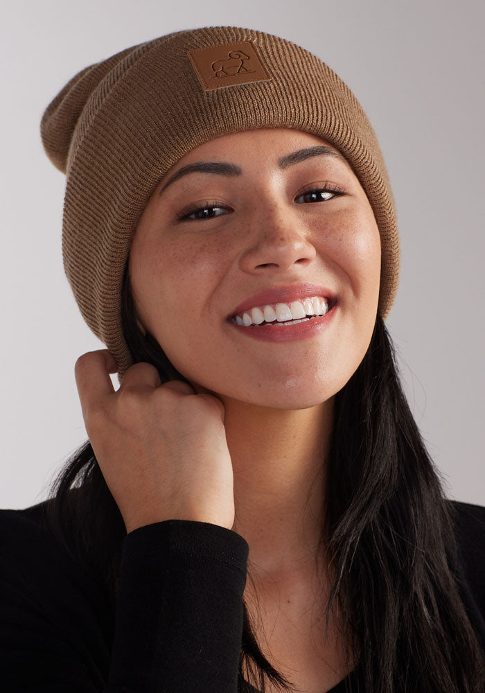 Wearing the cozy Baylor Beanie in camel by Woolx, a person with long dark hair smiles against a plain grey background while wearing a black top.
